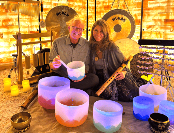 Two people smiling while seated among singing bowls, gongs, chimes, and other sound therapy instruments.