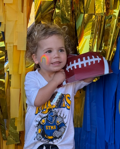 Little girl smiling holding a football in front of a blue and gold backdrop.