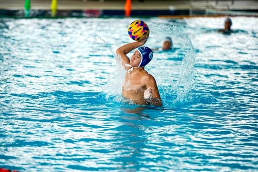 Athlete in water polo match splashing in indoor pool setting.