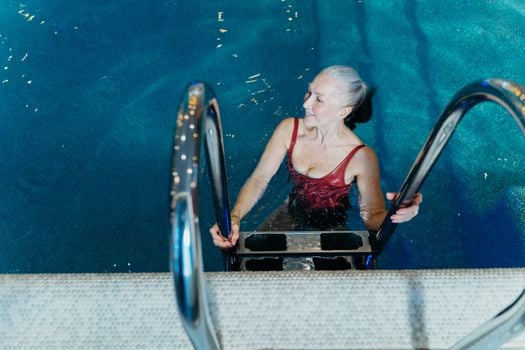 Elderly woman in a red swimsuit smiling while exiting a swimming pool.