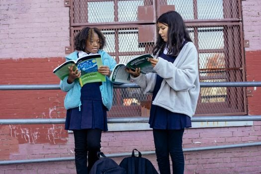 Focused diverse pupils helping each other doing homework with textbooks while standing near school